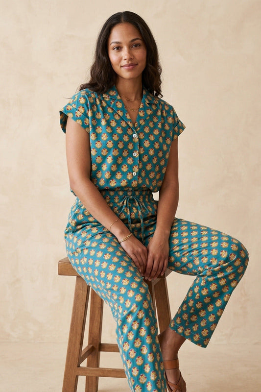 A woman sits on a wooden stool wearing a teal and yellow floral print matching set consisting of a short-sleeve button-front top and wide-leg pants, against a textured beige studio backdrop.