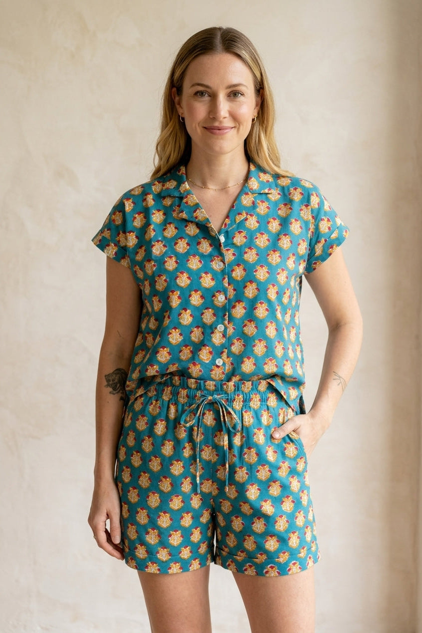 A woman modeling a matching short-sleeve button-front top and drawstring shorts set in teal with a repeating tan and cream floral pattern, standing against a warm beige textured wall.