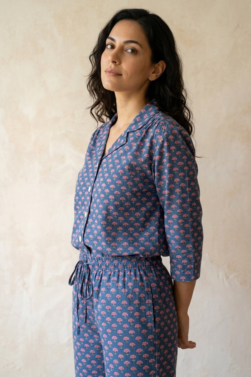 A woman wearing a blue and pink patterned matching set consisting of a button-up shirt and drawstring pants, standing against a textured beige wall.