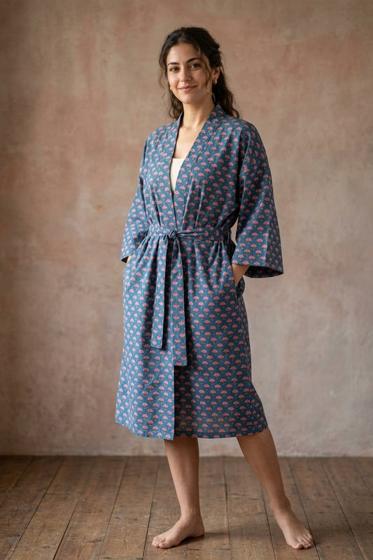 A woman wearing a blue cotton robe with a red and orange floral pattern, standing barefoot on a wooden floor against a textured pinkish-brown wall.