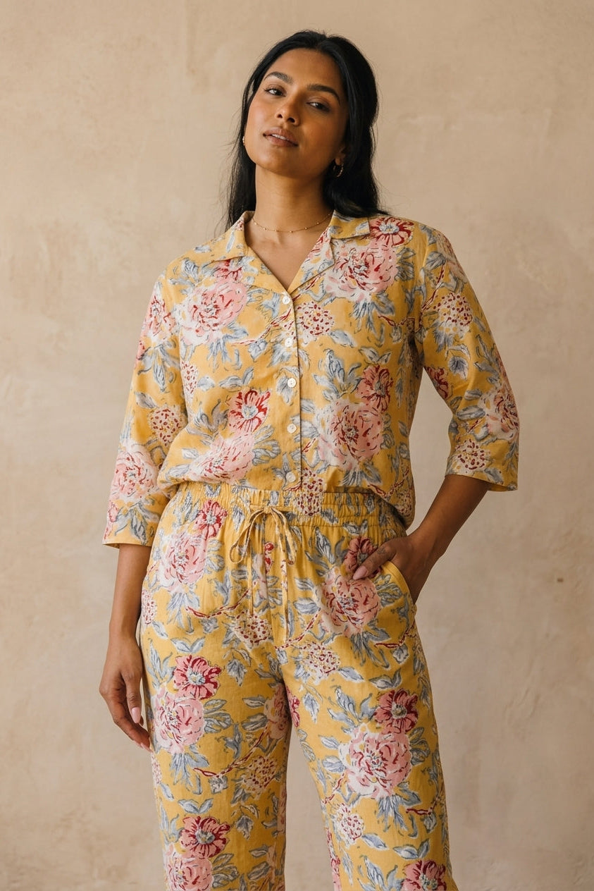 A woman modeling a matching two-piece pajama set with a yellow base and a pink and blue floral print, standing against a textured beige wall.