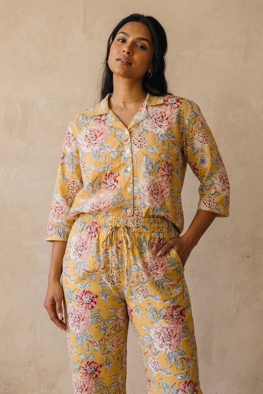 A woman modeling a matching two-piece pajama set with a yellow base and a pink and blue floral print, standing against a textured beige wall.