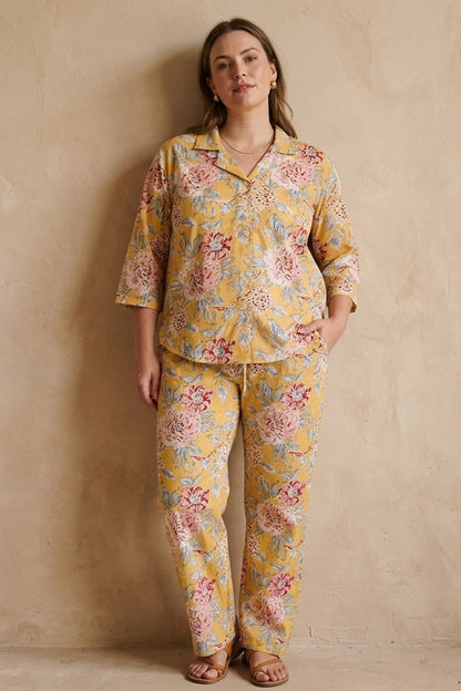 A woman stands against a textured beige plaster wall wearing a matching two-piece pajama set with a yellow base and a multicolor floral print in pink, red, and green.