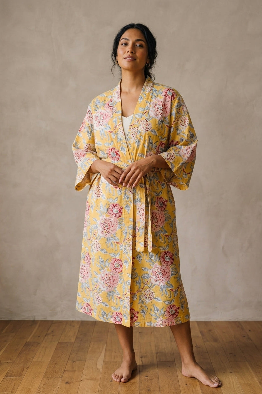 A woman stands barefoot in a studio wearing a yellow floral kimono-style robe with pink and blue flowers, against a textured beige wall and wood floor.
