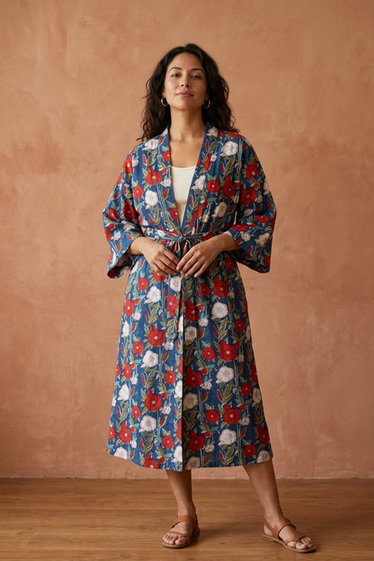 A woman stands against a warm terracotta wall wearing a long-sleeved floral robe with a blue base and red, white, and green botanical print, paired with tan sandals.